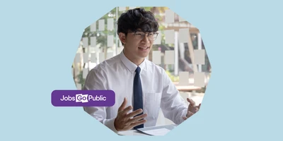 A man in glasses, a shirt, and tie is sat at a desk speaking animatedly to a hiring manager off screen