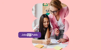 A female public sector teacher in a state school is leaning over a desk helping a female student with her work