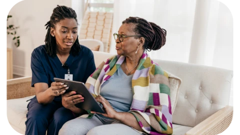 A female care worker in scrubs is sitting on a couch with a woman in her 70s, teaching her how to use a tablet device