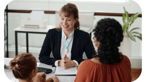 A female social worker in a blazer, shirt and lanyard is sitting at a desk opposite a little girl and foster carer whose backs are to the camera
