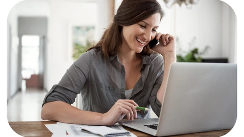 A female administrative assistant is sat in front of a laptop smiling and speaking to someone on a smart phone
