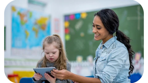 A female teaching assistant in SEND is working with a young girl with Down's syndrome, teaching her how to use a tablet