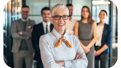 A female manager in glasses is standing mid-frame with her arms crossed and smiling. A team of 5 staff are standing behind her in business attire, slightly out of focus.