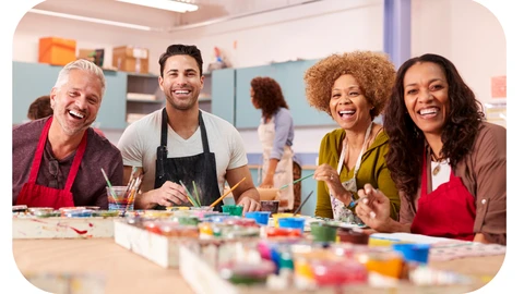 A diverse group of adults are sitting around a table at a community centre with painting tools