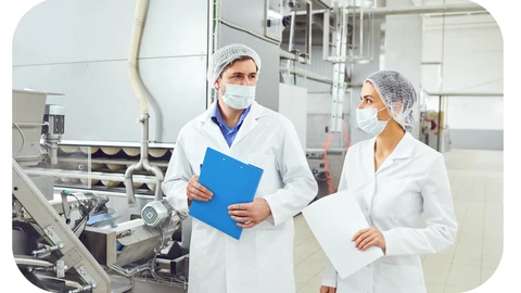 Two environmental health inspectors in lab coats, face masks, and hair nets are walking through a factory doing an inspection
