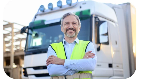 An HGV driver in the transport occupation is stood in front of a lorry in a high visibility vest and smiling with his arms crossed